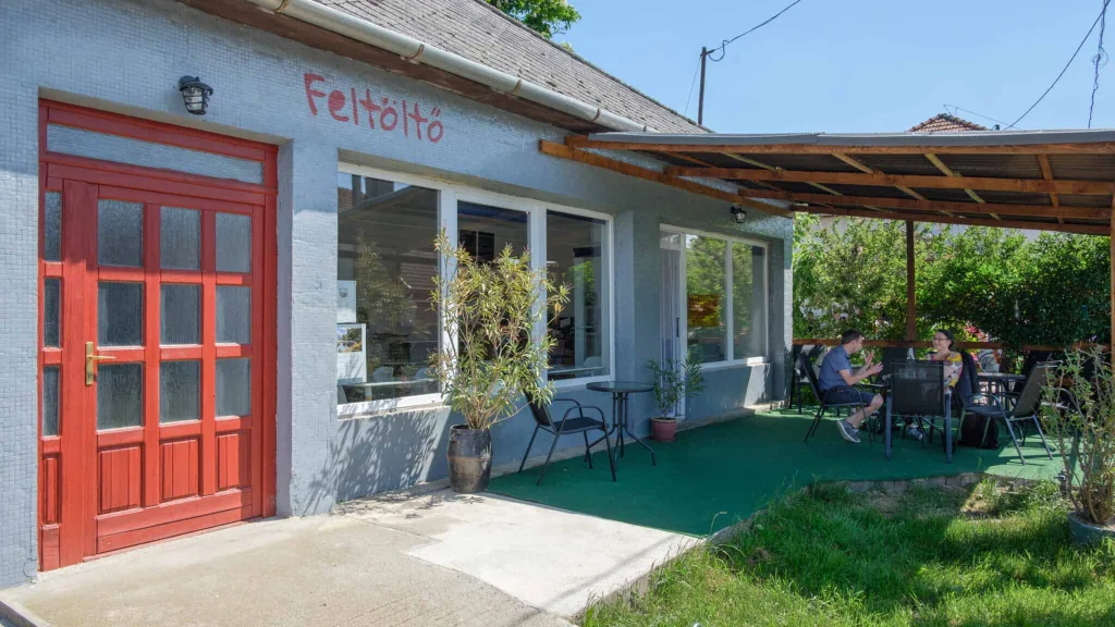 A coffeehouse in Hungary. A grey building with large windows and a red door. "Feltolto" is written on the wall in red. Customers are seen sitting under a verandah outside drinking coffee.