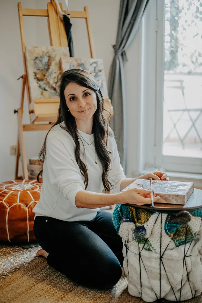 A female artist is seen kneeling on the floor working on a cavas. She looks at the camera reflectively. Other artworks are visible in the background.