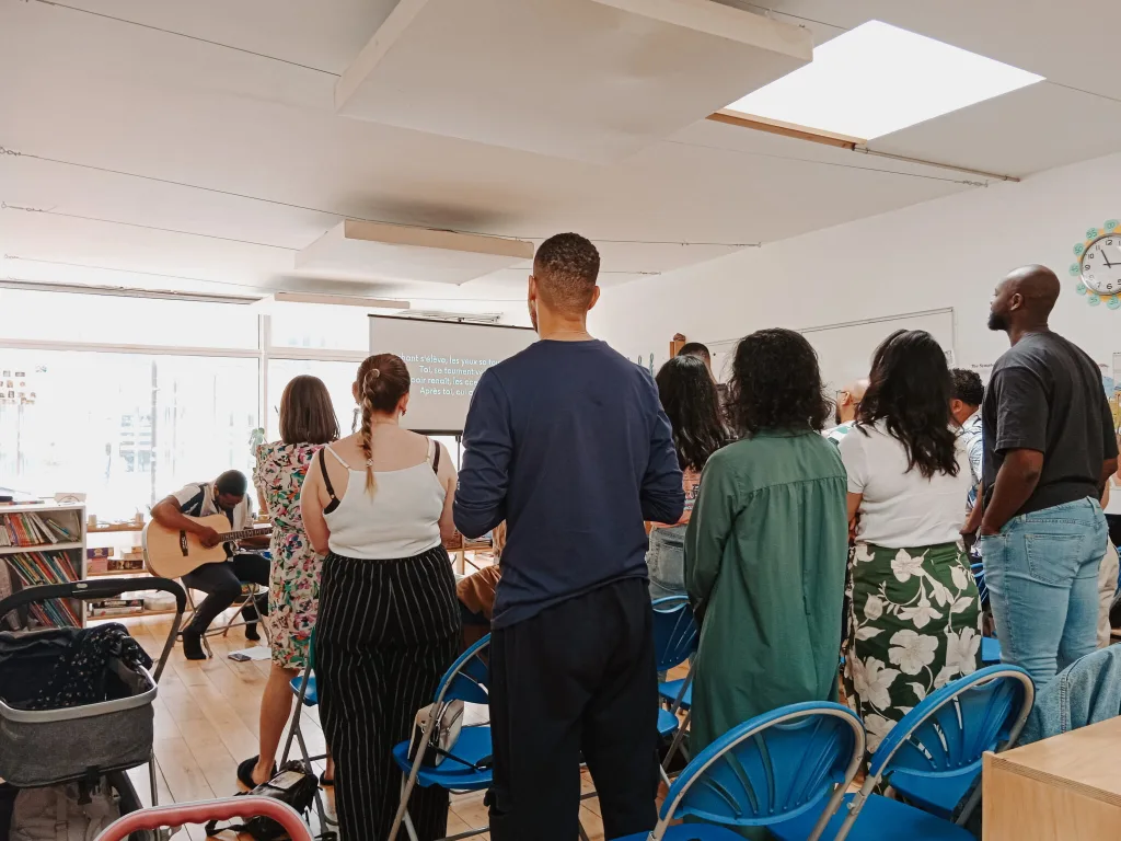 A group of adults of all ages sitting in rows in a school classroom. There is a man playing a guitar and worship lyrics on the screen at the front.